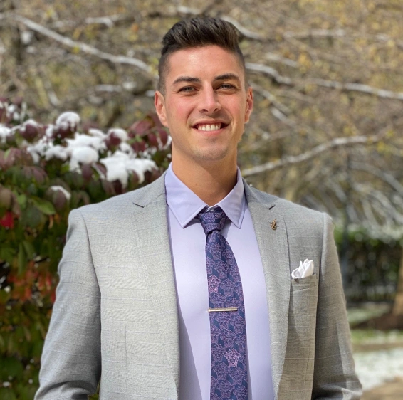 Professional man in business attire smiling outdoors with snowy evergreen background.