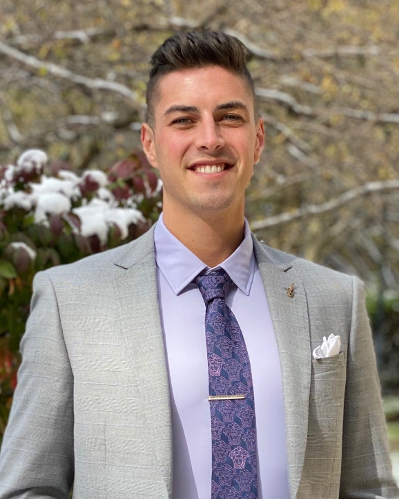 Professional man in gray suit and purple tie smiling outdoors with snow-covered foliage background.
