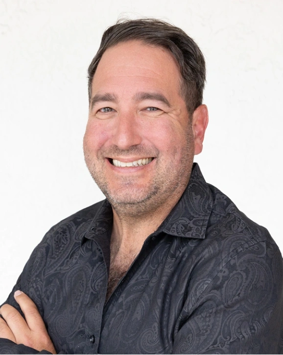 Professional headshot of smiling male dental specialist in dark shirt against white background.